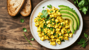 A vibrant tofu scramble served on a white plate, garnished with fresh parsley, alongside toasted bread and avocado slices on a rustic wooden table.